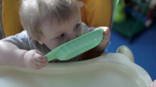 Een klein kind zit aan een hoge kindertafel en likt een bord. — Stockvideo