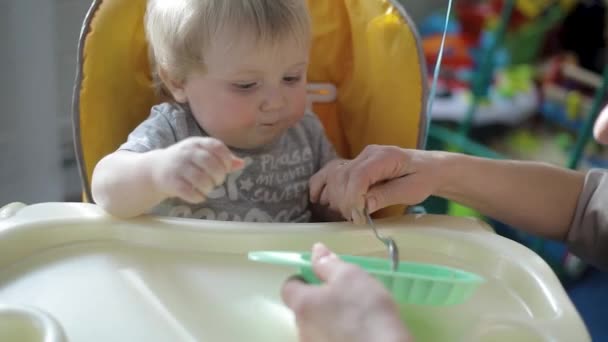 Grootmoeder voedt liefdevol haar kleinzoon, de baby zit op een kinderstoel. — Stockvideo