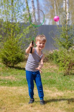 Sarışın bir çocuk çimenlerde badbinton oynar.