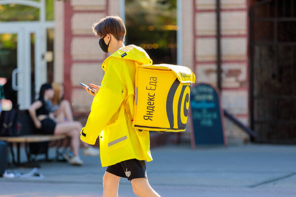 A student in a yellow jacket and a backpack delivers food, looks at the phone