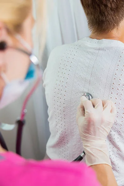 A girl doctor in a mask listens with a stethoscope to the back of an adult woman. — Stock Photo, Image