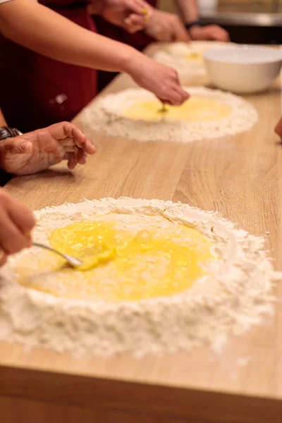 The hands of several people are kneading the dough on the table. Mix a raw egg with flour.
