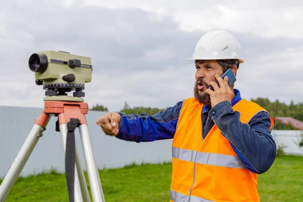 A disgruntled civil engineer with an optical level gauge speaks on a cell phone and swears.