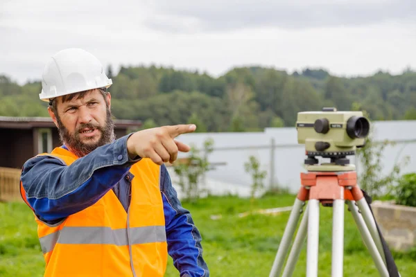 Stavební inženýr s optickou úrovní mávne rukou, aby ovládl kolegy z dálky. — Stock fotografie