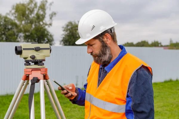 Stavební Inženýr Optickou Úrovní Podívá Mobilního Telefonu Vousatý Muž Hledá — Stock fotografie