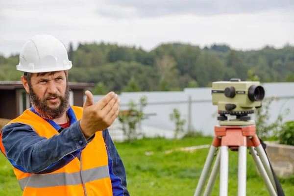 A civil engineer with an optical level waves his hand to control colleagues from a distance. A bearded man leads his subordinates.