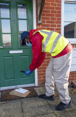 Hampshire, England, UK. 2020, Male courier delivering parcels and packages during Covid-19 epidemic wearing gloves and a mask. Recording on mobile phone the delivery. clipart