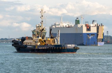 Southampton Water Southampton İngiltere. 28.09.2025. Svitzer Mercurius, İngiltere 'nin güneyindeki Southampton Water' ın arka planında bir araba taşıyıcısı ile römorkörü çekiyor.