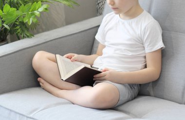 Kanepede kitap okuyan çocuk bacak bacak üstüne attı. Gri ve beyaz açık renkli fotoğraf. Yakın plan, görünürde yüz yok.