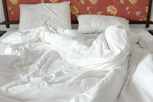 Unmade / untidy bed with a white crumpled blanket and two messy pillows in a bed room.