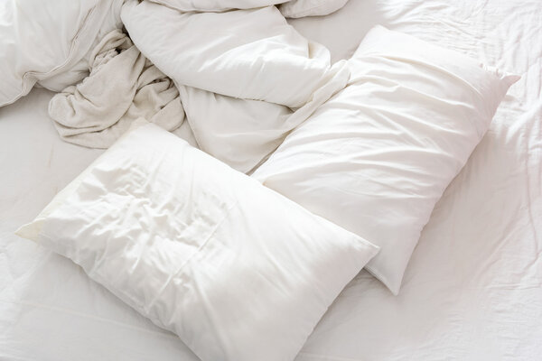 Top view of an unmade bed in a bedroom with crumpled bed sheet, a blanket, a white shower towel and two pillows.