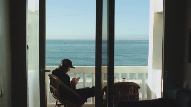 Joven disfrutando de la vista al mar desde la terraza. Movimiento lento Viajero masculino mirando el teléfono en el balcón con hermoso océano Taghazout Marruecos, 4k — Vídeo de stock