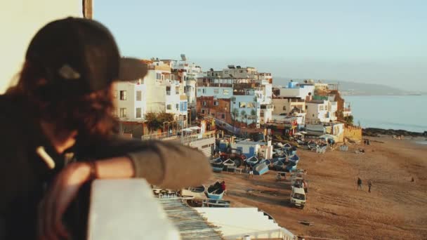 O homem na varanda está a olhar para a cidade. Movimento lento. Bela aldeia e pequena praia aconchegante Oceano Atlântico em Marrocos, 4k — Vídeo de Stock