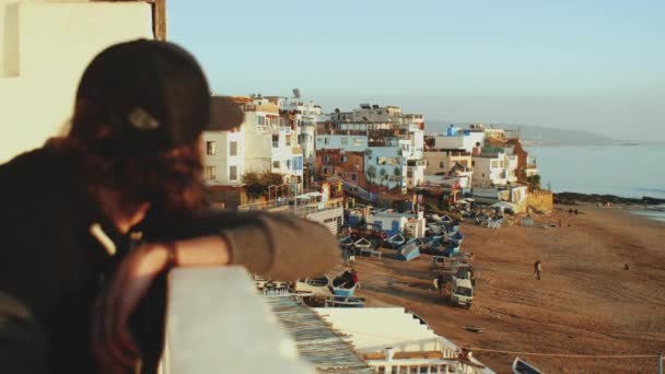 De man op het balkon kijkt naar de stad. Langzame beweging. Mooi dorp en klein gezellig strand Atlantische Oceaan in Marokko, 4k — Stockvideo