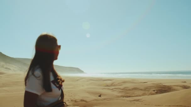 Een vrouw met een zonnebril kijkt naar de oceaanhorizon. Amazing Sand Dunes by Beautiful Blue Ocean, Dahkla, Marokko, zonnekonijnen in de camera, Slow motion, 4k — Stockvideo