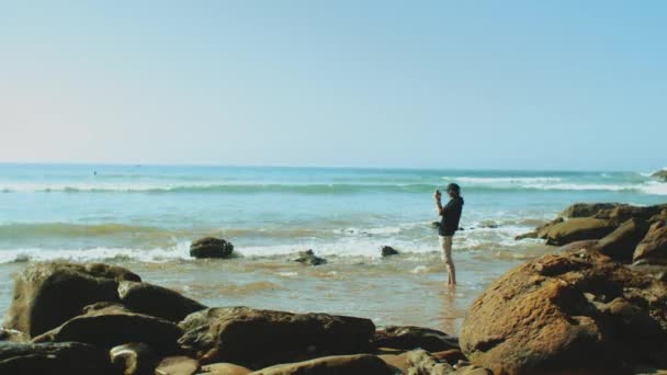 Al rallentatore. Uomo scattare foto su smartphone sulla spiaggia, l'oceano sullo sfondo, costa sassosa, turista in Marocco, 4k — Video Stock