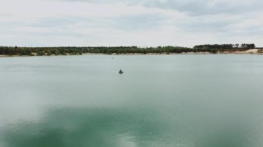 Gölün ortasında teknede yalnız bir adam, çam ağacı ormanıyla yaz mevsiminde doğal göl manzarası. Ukrayna doğası, güzel göl ve yeşil orman üzerindeki hava.