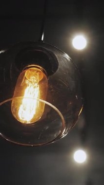 Dikey video. Siyah tavan zemininde asılı yuvarlak avize. Abajur ve klasik ampul, top lambası. Yakın plan çekim. Eski çatı katı lambaları. Yüksek kalite 4k görüntü