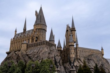 NOV 27 2025 Evrensel Stüdyoları Japonya: Hogwarts Şatosu yemyeşil ve kayalık arazilerle çevrili bir tema parkı ortamında bulutlu bir gökyüzüne karşı görkemli bir şekilde duruyor