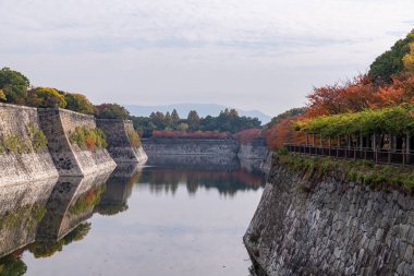 Osaka, Japonya 'daki bir şehir parkının arka planında sonbahar yaprakları, dağlar ve ağaçlarla dolu sakin bir suyolu çevreleyen tarihi taş duvarların görüntüsü - 24 Kasım 2025