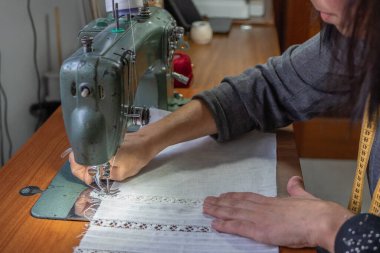 Kadının elleri eski bir dikiş makinesiyle beyaz kumaş dikiyor.