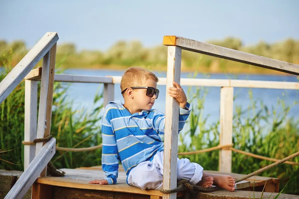 Bellissimo ragazzo in posa seduto su un ponte di legno. Vestita con un tema nautico  . — Foto Stock