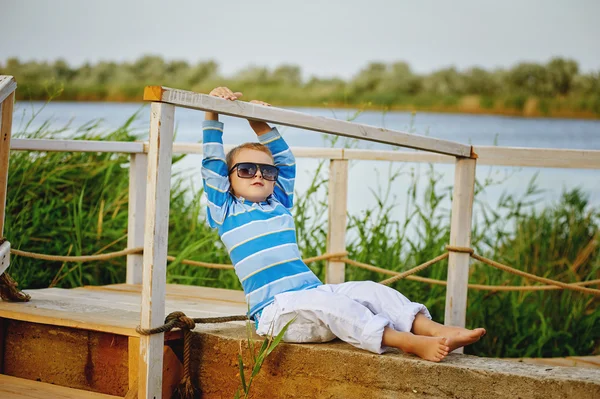 Bellissimo ragazzo in posa seduto su un ponte di legno. Vestita con un tema nautico  . — Foto Stock