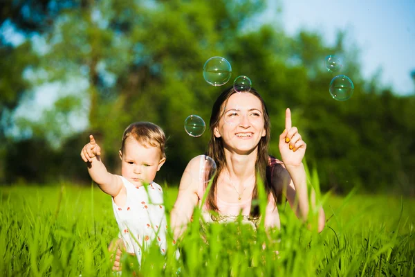 Mom and baby girl laying on grass and cheerfully catching soap b — Stock Photo, Image