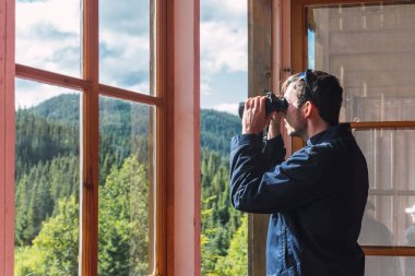 Dürbünden ormana bakan bir adam ahşap bir kulübeden ormana bakıyor.