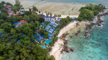 Lüks bir ada tatil köyünün mavi çatılı kulübeleri ve kumsalda yüzme havuzu olan verimli bir ormanın yüksek açılı görüntüsü. Seyahat acentesinin broşürleri ve konukseverlik pazarlaması için ideal. Yüksek kalite fotoğraf