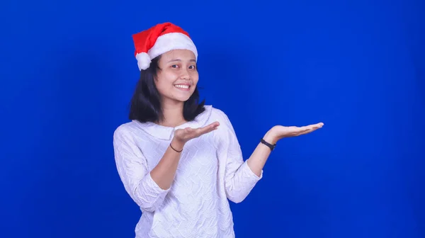 右の空白を指してクリスマスの帽子をかぶっているアジアの女性隔離された青の背景 — ストック写真