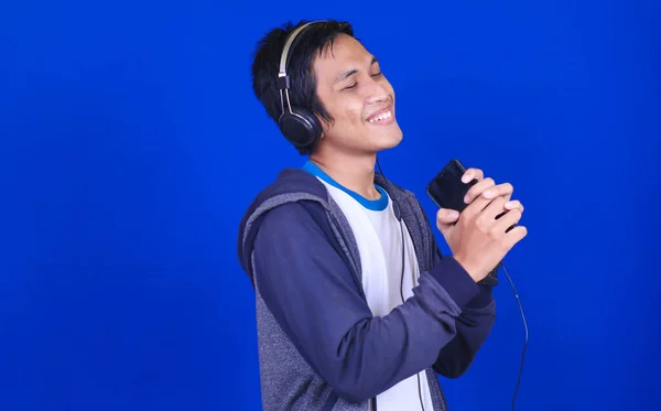 亚洲男人带着快乐的表情听着音乐 戴着手机和耳机 背景是蓝色的 — 图库照片