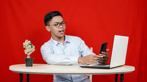 Wow face of Young Asian man shocked and surprised what he see in the smartphone and laptop on the table