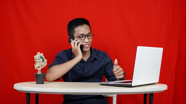亚洲商人打电话来 桌上拿着笔记本电脑 背景是孤立的红色 — 图库照片
