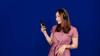Asyalı kadın, mavi arka planda kulaklık kullanarak şarkı dinliyor.
