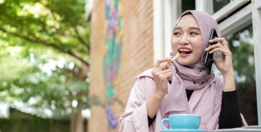 Mutlu Asyalı tesettürlü kadın akıllı telefon kullanıyor ve arıyor, kafede otururken kahve içiyor.