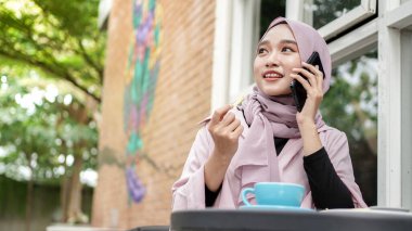 Mutlu Asyalı tesettürlü kadın akıllı telefon kullanıyor ve arıyor, kafede otururken kahve içiyor.