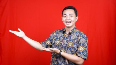 Batik giyen Asyalı bir adam boş alanı işaret ederek izole edilmiş kırmızı arka plan