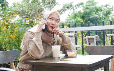 Mutlu Asya tesettürlü kadın akıllı telefon kullanıyor, arıyor, kafede oturuyor.