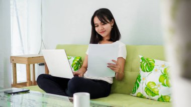 Genç, güzel, Asyalı bir kadın evde dizüstü bilgisayar kullanıyor ve sandalyede oturuyor.