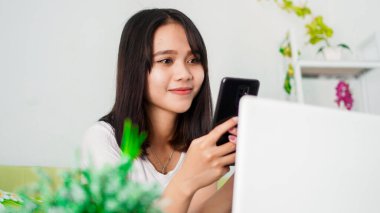 Asyalı kadın evde çalışırken koltukta dizüstü bilgisayarla cep telefonu kullanıyor.