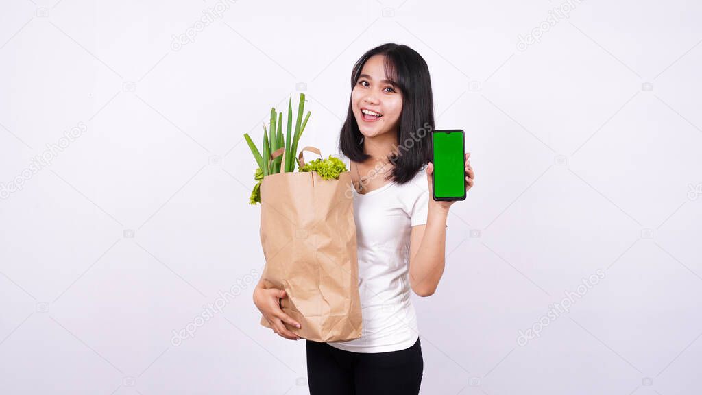 Asian woman with paper bag of fresh vegetables and holding a green screen phone with isolated white background