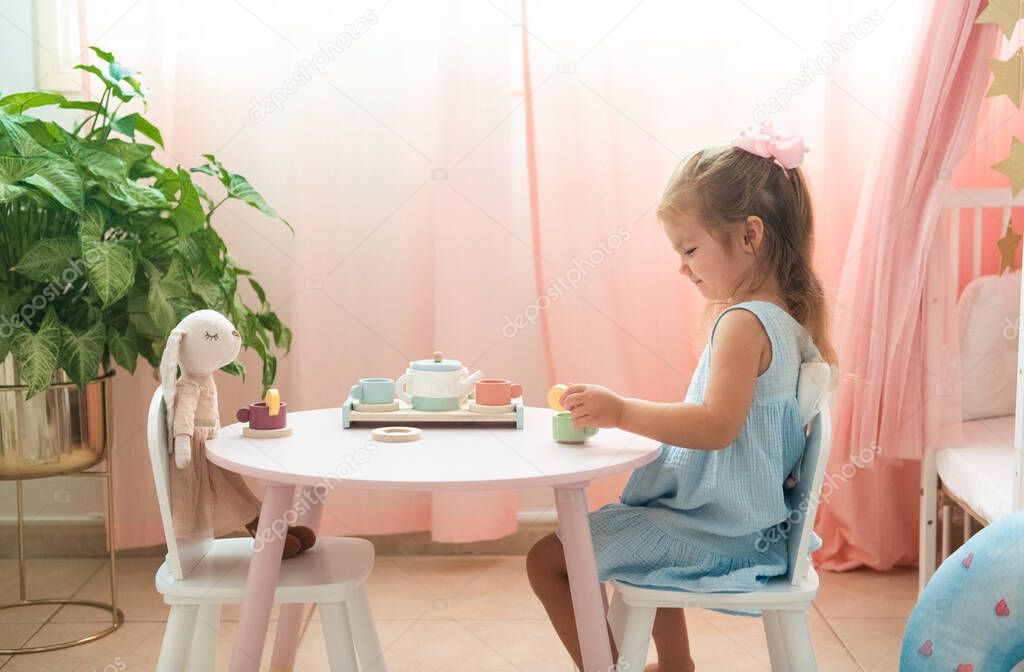 Toddler caucasian fair haired girl in blue dress is role playing drinking tea with rabbit toy in her pink beautiful room
