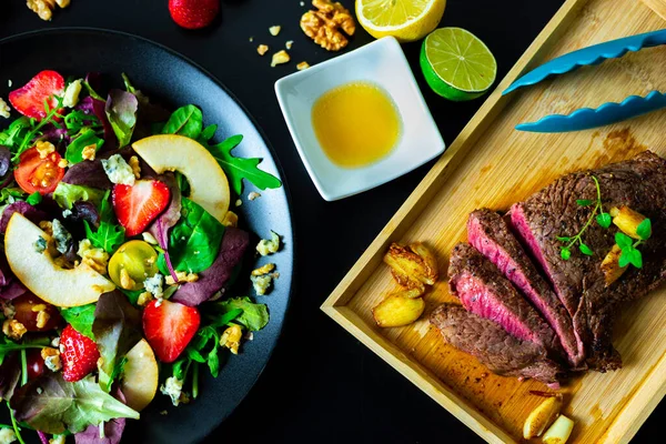 Cut steak on wooden dish with side of mixed salad with strawberries, pears, walnuts, blue cheese and lemon-honey dressing