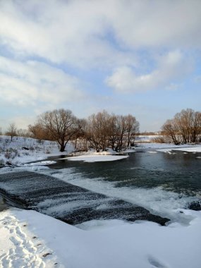 Küçük suyu olan nehir barajı dökülüyor ve mavi bulutlu gökyüzü altında kıyılar karla kaplanıyor