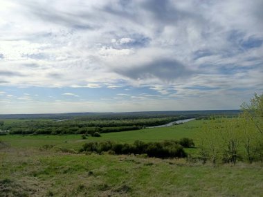 Mavi gökyüzünün altındaki iki nehir arasında çimenli ve ağaçlı yeşil tarlalar