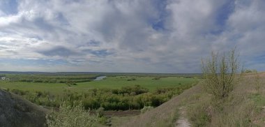 Mavi gökyüzünün altındaki iki nehir arasında çimenli ve ağaçlı yeşil tarlalar