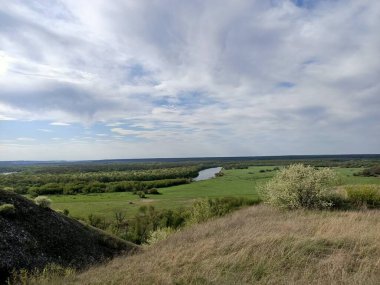 Mavi gökyüzünün altındaki iki nehir arasında çimenli ve ağaçlı yeşil tarlalar