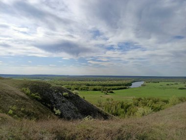 Mavi gökyüzünün altındaki iki nehir arasında çimenli ve ağaçlı yeşil tarlalar
