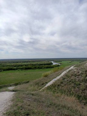 Mavi gökyüzünün altındaki iki nehir arasında çimenli ve ağaçlı yeşil tarlalar